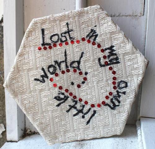 Hexagon shaped fabric patch with &quot;lost in my own little world&quot; stitched in black thread, spiralling into the centre.